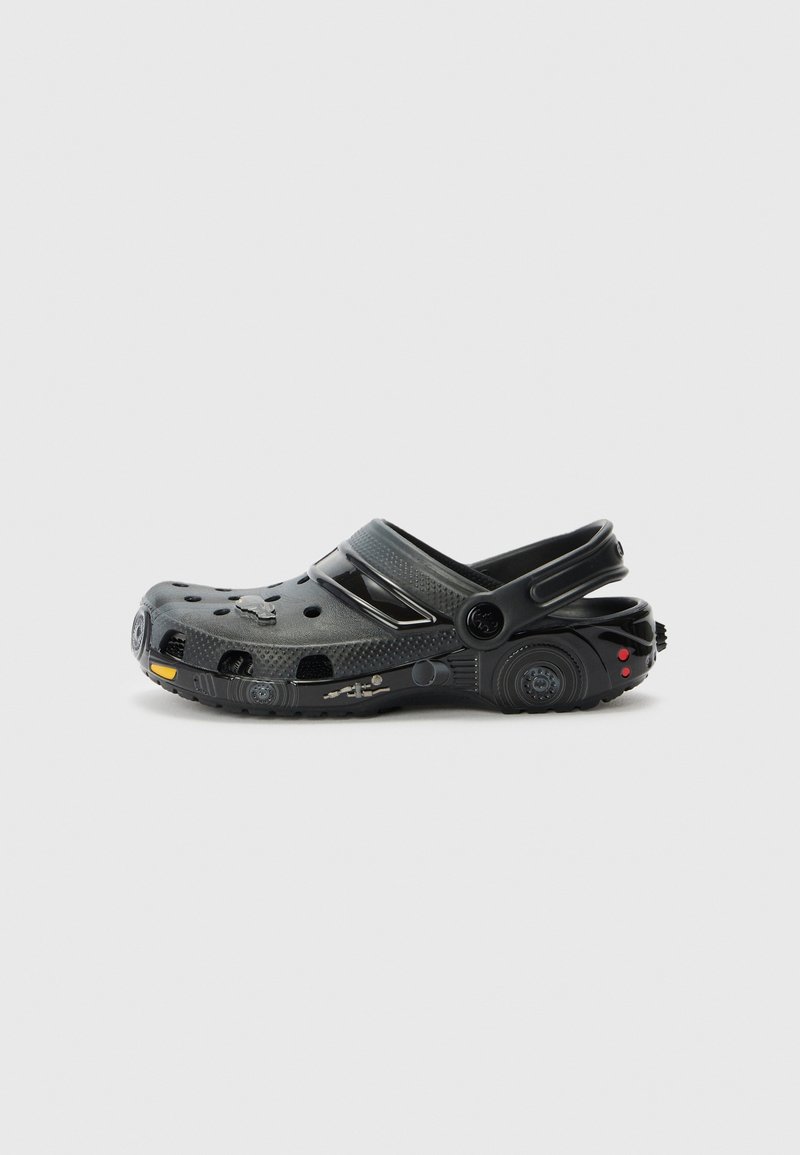 Chaussure style sabot noire avec des trous de ventilation et une bride arrière, présentant des éléments de design détaillés ressemblant à un véhicule sur la semelle et le talon.