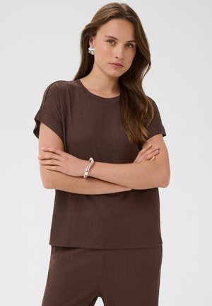 Femme aux longs cheveux bruns portant un haut marron à manches courtes et un pantalon assorti, des boucles d'oreilles argentées et un bracelet argenté, les bras croisés.