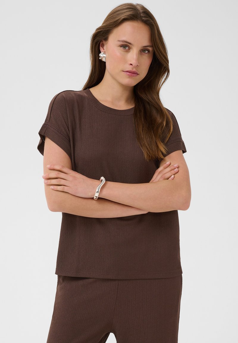 Femme aux longs cheveux bruns portant un haut marron à manches courtes et un pantalon assorti, des boucles d'oreilles argentées et un bracelet argenté, les bras croisés.