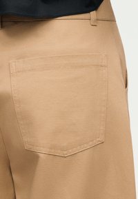 Κοντινή λήψη μπεζ παντελονιού που δείχνει πίσω τσέπη με ορατό ράψιμο και μαύρο πουκάμισο κρυμμένο στη μέση.