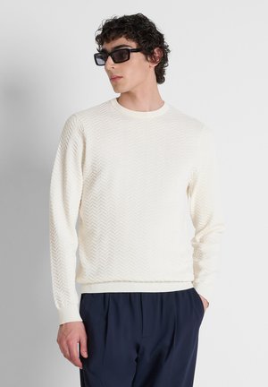 Modello maschile che indossa un maglione bianco a maniche lunghe con trama, pantaloni blu scuro e occhiali da sole rettangolari neri, posando con una mano in tasca.