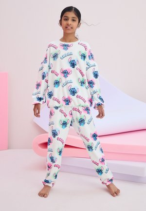 Dziewczynka stoi boso, ubrana w białą piżamę z kolorową kreskówką z Stitch i napisem "Ohana" na pastelowym różowym tle.
