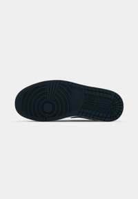 Semelle en caoutchouc noire d'une sneaker montrant des motifs de bande de roulement circulaires et linéaires pour l'adhérence et la traction, photographiée sur fond blanc.