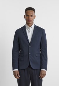 Sötétkék blézer, szabott fazonnal, két elülső zsebbel, egygombos záródással és finom textúrájú anyagból, csíkos ing felett viselve.