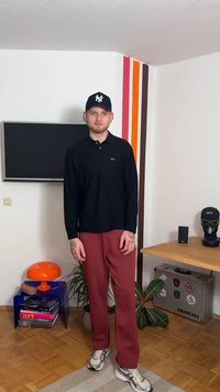 Polo noir à manches longues avec un logo doré, associé à un pantalon large rouge et des baskets grises. En arrière-plan, des rayures murales colorées et du mobilier.