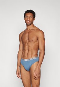 Blaue Baumwollslips mit einem grauen gestreiften Bund, die eine enganliegende Passform und seitliche Einschnitte aufweisen. Das Modell steht leicht nach rechts gewandt.