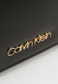 Černá texturovaná kožená taška s zlatým logem "Calvin Klein". Materiál je jemný s výraznou strukturou a elegantním, minimalistickým designem.