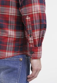 Poignet de chemise à manches longues à carreaux rouge et marine avec des boutons noirs, portée avec un jean en denim bleu montrant la poche arrière et une main détendue le long du corps.