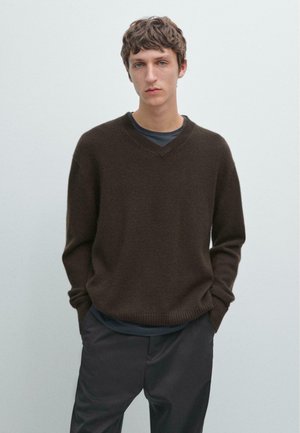 Pull en maille brun avec un col en V et des poignets côtelés. Porté sur un haut sombre, associé à un pantalon foncé. Design simple et décontracté.