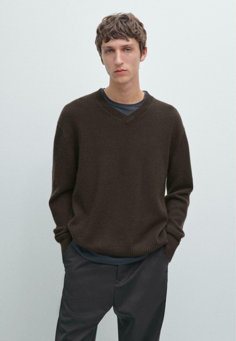 Maglione marrone texture con scollatura a V e polsini a costine. Indossato sopra una maglietta scura, abbinato a pantaloni scuri. Design semplice e rilassato.