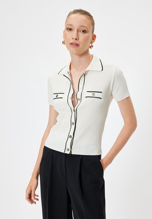 Kurzärmliges, weißes geripptes Shirt mit kragen, schwarzen Paspelierungen und zwei Brusttaschen mit runden Knöpfen.
