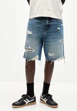 Short en denim avec plusieurs déchirures effilochées, porté avec des chaussettes noires et des baskets noires avec des bandes blanches et des semelles en gomme.