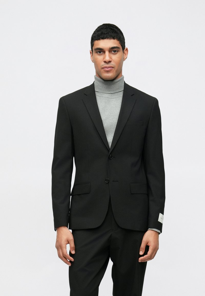 Μαύρο σακάκι με δύο κουμπιά, με κανονικούς γιακάδες και πλαϊνές τσέπες. Φοριέται πάνω από ένα ανοιχτό γκρι πουλόβερ με ζιβάγκο. Λείο φινίρισμα υφάσματος.