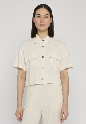 Donna con capelli lisci e scuri che indossa una camicia beige chiaro a maniche corte con tasche sul petto e pantaloni abbinati.