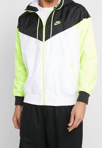 Nike vindjacka med svart överdel, vit kropp och neongröna ärmar. Inkluderar ficka med dragkedja och justerbar huva.