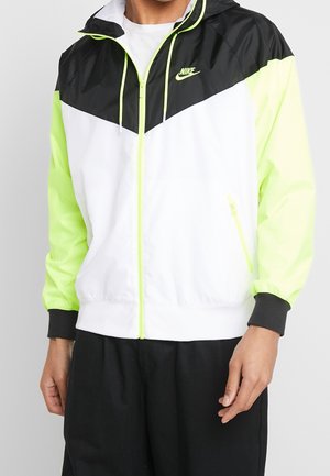 Kurtka przeciwwiatrowa Nike z czarną górą, białym korpusem i neonowozielonymi rękawami. Zawiera zapinaną na zamek kieszeń oraz regulowany kaptur.
