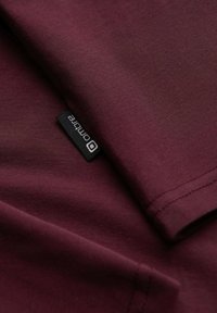 Burgundyröd bomullströja med en mjuk textur, med en sydd svart etikett som visar varumärket, delvis vikt vid kragen.