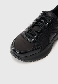 Chaussure de sport noire avec un dessus en synthétique, des accents en maille, des détails brillants et une semelle texturée. Les lacets sont noirs et la chaussure est dotée d'un collier rembourré pour le confort.