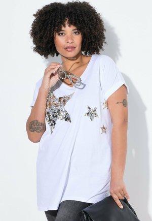 Bílý T-shirt s výstřihem do V, na přední straně zdobený zlatými a stříbrnými flitry ve tvaru hvězdy. Krátké rukávy a volný střih, spárovaný se šedými džíny.