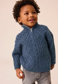 Maglione lavorato a maglia blu con colletto alto, caratterizzato da texture a trecce e una zip a quarto. Abbinato a jeans grigio chiaro.