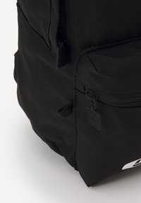 Sac à dos noir en tissu avec fermetures éclair et détail de logo, présentant une surface texturée et une forme structurée pour la durabilité et l'utilité.