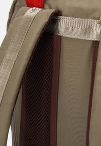 Khaki backpack with a textured mesh panel, reinforced brown trim, and silver straps. Features a red accent at the top.