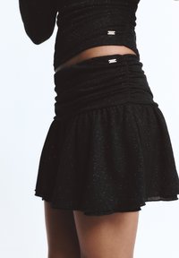 Fekete szoknya bővülő fazonnal és húzott derékkal. Az anyag finoman csillog, fémes díszítéssel a derékvonalnál.