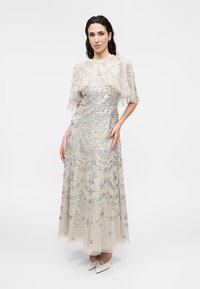 Γυναίκα που φοράει μακρύ μπεζ φόρεμα με ασημένιες παγιέτες και floral σχέδια, διαφανή κοντά βολάν μανίκια, στέκεται με το χέρι στη μέση.