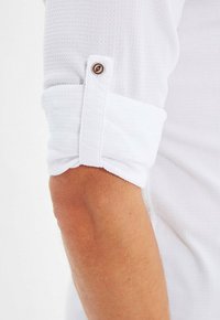 Biała koszula z długim rękawem wykonana z teksturowanej tkaniny, mankiet podwinięty i zapinany na brązowy guzik, prezentująca czysty, minimalistyczny design.