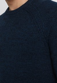 Man som bär en mörkblå strukturerad stickad tröja med synliga sömdetaljer runt axlar och halsringning.