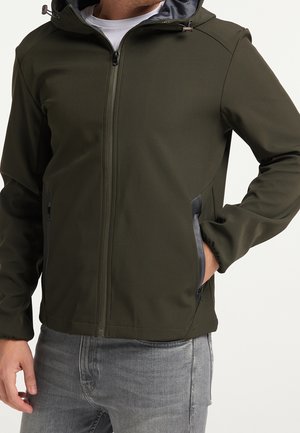 Homme portant une veste à capuche vert olive avec fermeture éclair à l’avant et poches latérales, associée à un jean gris clair.