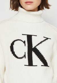 Pull à col roulé crème en maille avec un grand logo "CK" noir sur le devant, présenté sur un tissu texturé avec des détails côtelés.