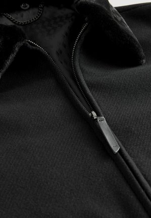 Schwarze Fleecejacke mit Reißverschluss, strukturiertem Stoff und einem Kragen mit weichem Besatz. Innen zeigt ein gemustertes Futter.