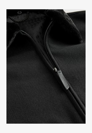 Schwarze Fleecejacke mit Reißverschluss, strukturiertem Stoff und einem Kragen mit weichem Besatz. Innen zeigt ein gemustertes Futter.