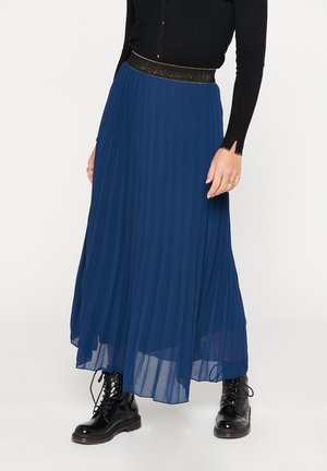Femme portant un haut noir à manches longues, une jupe longue plissée bleue avec une ceinture scintillante, et des bottes de combat noires à lacets.