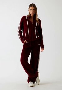 Trening din catifea vișinie cu jachetă cu fermoar și dungi albe, completat de pantaloni asortați. Ținuta este întregită de adidași albi.