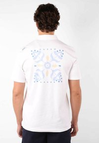 T-shirt en coton blanc avec un grand motif floral bleu et jaune clair au dos. Manches courtes et col rond.