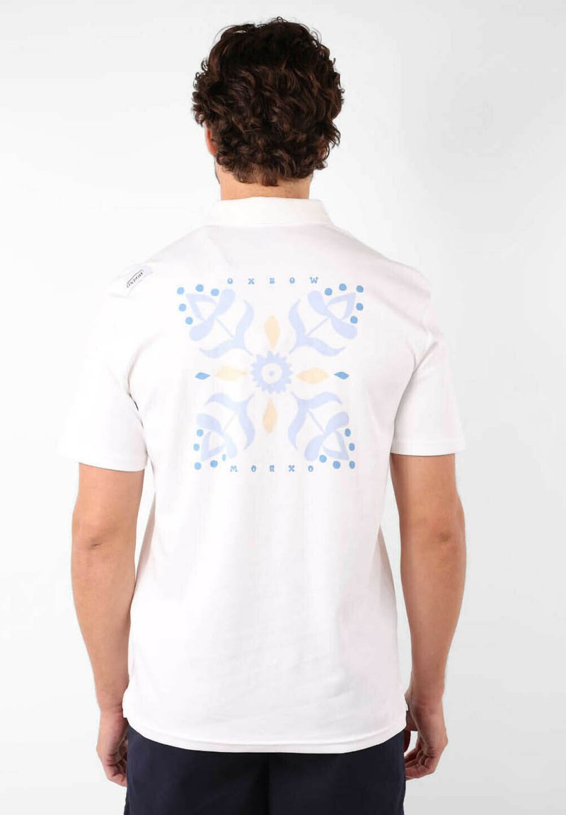 T-shirt en coton blanc avec un grand motif floral bleu et jaune clair au dos. Manches courtes et col rond.