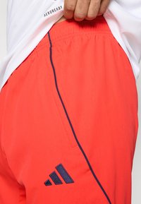 Czerwone sportowe spodenki z teksturowaną powierzchnią. Posiadają granatowe boczne przeszycia oraz logo Adidas w kolorze granatowym na dole po lewej stronie. Pokazany jest elastyczny pas.