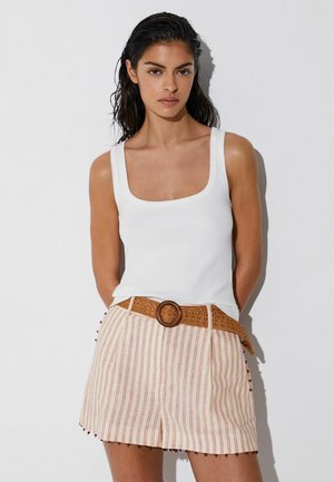 Nő nedves hajjal, fehér ujjatlan felsőt és bézs csíkos rövidnadrágot visel fonott övvel, egyszerű fehér háttér előtt.