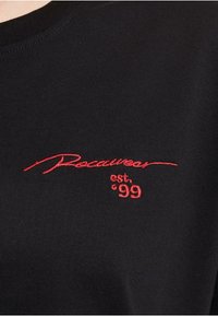 Black t-shirt featuring a ribbed texture and round neckline, with red embroidered logo that reads "Rocawear est. '99" on the chest.