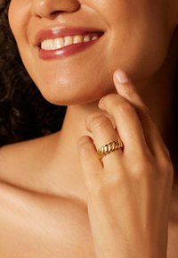 Gouden ring met een golvende bandontwerp met afwisselend gladde en gestructureerde segmenten, gedragen op een hand die bij het gezicht rust.
