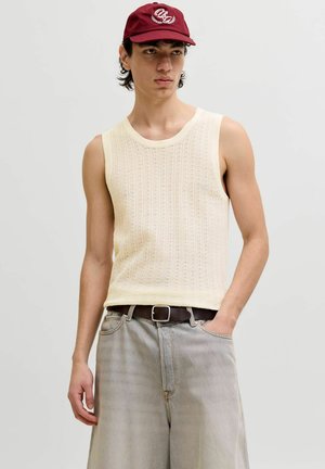 Jeune homme portant un haut en maille crème sans manches, un jean gris clair, une ceinture marron et une casquette de baseball rouge, debout avec une main dans la poche.