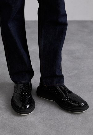 Chaussures en cuir noir avec un motif perforé, bout rond et design à lacets, associées à un jean denim foncé avec des coutures orange.