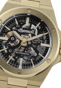 Goudkleurig automatisch horloge met een skeleton-dial, zwarte accenten en zilveren uurmarkeringen. Geborstelde metalen band met hexagonale schroeven.