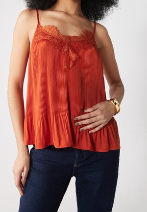 Femme portant un haut sans manches plissé orange avec bordure en dentelle, jeans bleu foncé, bracelet doré, main posée sur la taille.