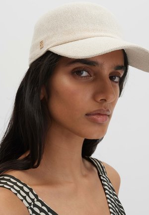 Femme aux longs cheveux foncés portant une casquette écrue texturée et un haut sans manches à motif noir et blanc, légèrement de profil.