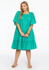 Teal katoenen tiered jurk met korte fladdermouwen en vierkante hals, met plooidetails en een comfortabele pasvorm, gedragen met tan sandalen.