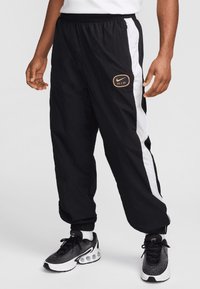 Pantalones de nailon negros con paneles laterales blancos y un logotipo de Nike Air en naranja. Cuentan con una cinturilla elástica y puños estrechos. Combinados con zapatillas negras.