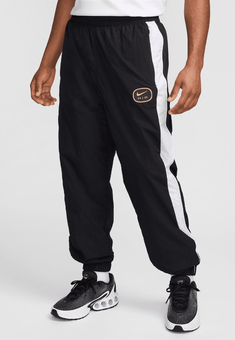 Pantalones de nailon negros con paneles laterales blancos y un logotipo de Nike Air en naranja. Cuentan con una cinturilla elástica y puños estrechos. Combinados con zapatillas negras.